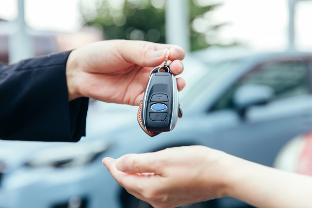 車のキーを手渡している手元のクローズアップ。背景に駐車中の車