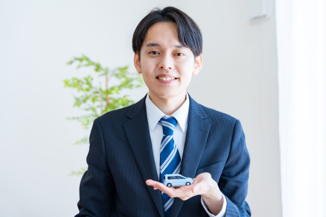 スーツ姿の男性が、手のひらに小さな車の模型をのせて差し出している様子